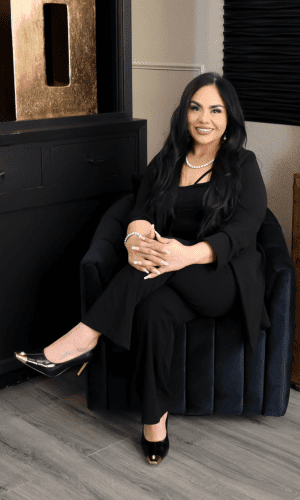 Woman in black sitting on a chair, smiling with hands clasped.